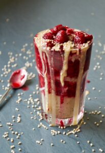 Ein Glas mit Porridge und heißen Himbeeren gefüllt, dazu Erdnussbutter. Daneben gepuffter Amaranth und ein Löffel